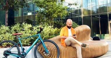 Şehir merkezinde bisiklet süren yakışıklı, sakallı, gülümseyen adam. Profesyonel genç erkek işe gidip telefonla yazarken arka planda bisikletle bankta oturuyor..