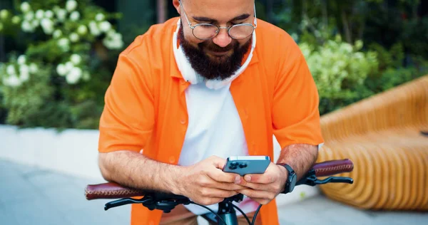 Handsome smiling man with bicycle wearing glasses texting on phone and looking around near building outdoors. Attractive happy male tapping and scrolling on mobile phone.