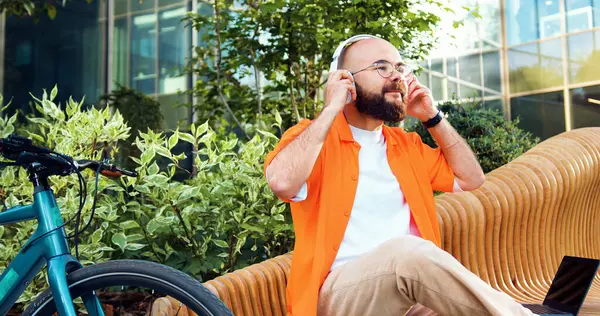 Cheerful bearded man in glasses wears headphones and listens to music outdoors. Hipster guy with bicycle using the phone sitting on bench near a modern office building.