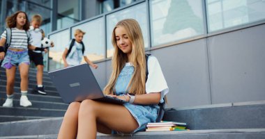 Genç ve güzel bir kız okul arkadaşlarını karşılamak için bilgisayar kullanıyor. Okul binasının yanındaki merdivenlerde oturuyor.