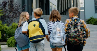 Group of four smart diverse classmates with colorful backpacks are having fun talking while going to school school, outdoors. Back view. clipart