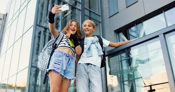 Okul binasının yanında kitap ve sırt çantalarıyla duran sevimli, komik, küçük bir kız ve erkek çocuk fotoğraf çekerken akıllı telefon kamerasına alaycı yüz ifadeleri takınıyorlar. Okul kavramı.