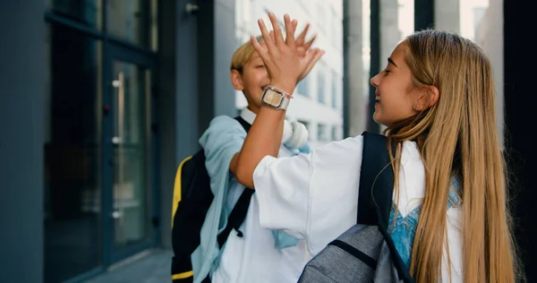 Genç öğrenciler okul yakınlarında buluşup selamlaşıyorlar, konuşuyorlar ve dışarıda beşlik çakıyorlar. Sırt çantaları ve laptoplarıyla okula giderken gülümseyen ve birbirini selamlayan pozitif okul çocukları.