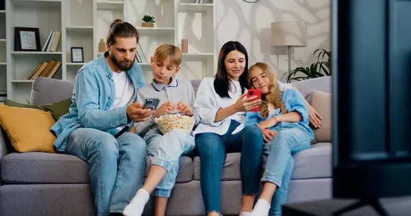 Çekici üzgün aile evdeki kanepede birlikte oturuyor ve herkes mesajları okumak ya da arkadaşlarıyla sohbet etmek için akıllı telefonu kullanıyor..