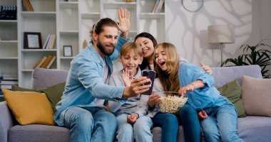 Mutlu aile bireyleri ve sevimli okul çocukları. Akıllı telefon uygulamaları kullanıyorlar. Evdeki kanepede oturup teknolojiyle video görüşmesi yapıyorlar. Anne, baba, anaokulu çocukları.
