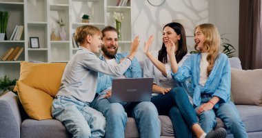 Mutlu, neşeli, genç aile kanepeye oturmuş dizüstü bilgisayardaki e-posta ile harika haberleri kutluyor, beşlik çakıyor ve birbirinizi başarıyla tebrik ediyor. Erkekler, kadınlar ve çocukların keyfi yerinde.