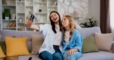Neşeli kız ve anne-baba evde telefonla komik fotoğraflar çekerken. Güzel gülümseyen bir aile kanepenin üzerinde akıllı telefon kamerasıyla selfie çektirir. Hafta sonu eğleniyor ve dinleniyoruz..