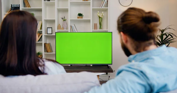 Rear View. Family couple watches green screen TV mockup sitting on couch in living room together. Man and woman watching TV chroma key in domestic cinema looking TV show or news in home.