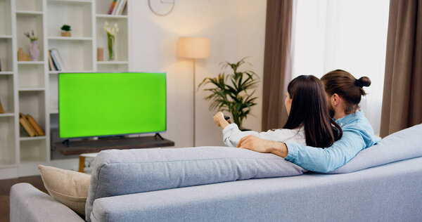 Caucasian happy couple man and woman having fun, sitting on sofa in living room watching at TV monitor with green chroma key screen. Woman switching channels with remote control. Resting at home in