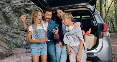 Güzel bir aile babası ve çocuklar bagajları açık bir şekilde arabada oturup gülüyorlar internette akıllı telefonlardan araklıyorlar. Mutlu bir aile. Oğulları ve kızlarıyla birlikte bir arabanın bagajında oturuyorlar.