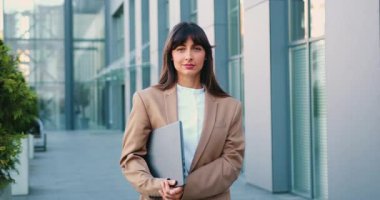 Uzun siyah saçlı profesyonel bir kadın dizüstü bilgisayar tutuyor ve son moda bir çalışma alanında duruyor. Açık havada, dostça bir gülümsemeyle kameraya bakıyor. Yavaş çekim