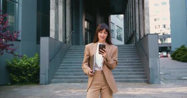 Şehirde bir iş gününden sonra modern bir ofis binasının önünde dikilirken resmi kıyafetli gülümseyen profesyonel bir kadın akıllı telefon kullanıyor. Yavaş çekim