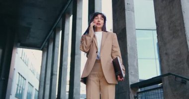 Modern bir ofis binasının önünde duran, elinde dizüstü bilgisayarla iş arkadaşlarıyla telefonda konuşan, iş gününün sonunda ciddi bir iş görüşmesi yapan kendine güvenen bir kadın. Yavaş çekim