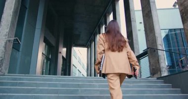 Kendine güvenen, uzun saçlı, resmi bir takım elbise giyen, bir elinde dizüstü bilgisayarla dışarıda yürüyen şehir ortamında modern ofis binasına yaklaşan bir iş kadını. Yavaş çekim