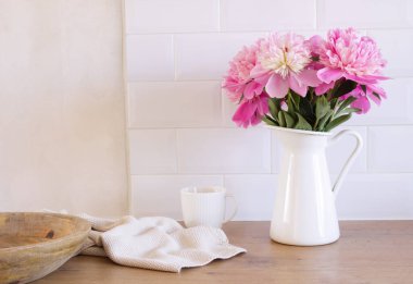 Peonies flowers in a vase and a cup of coffee on a wooden table. Blurred kitchen background contemporary elegant scandi interior.