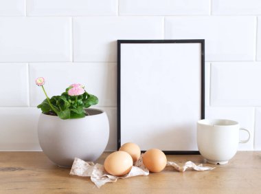 Bahar hala hayattır. Ahşap masa arkasında boş resim çerçevesi modeli. Leucanthemum vulgare, çanakta Bellis perennis çiçekleri, tavuk yumurtaları ve ipek kurdeleli Paskalya kompozisyonu. Çiftlik evi