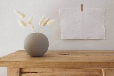 Beyaz bir duvara bantlanmış boş kağıt çarşaflar ve posterler. Tahta bank, masa. Kuru Lagurus ovatus çimi ve fincan kahvesiyle modern beyaz seramik vazo. İskandinav iç mimarisi. Seçici odak.