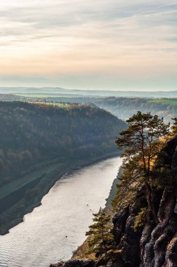 Su, yüksek dağlar ve yemyeşil ağaçlar tarafından kucaklanan bir vadiden akar ve açık gökyüzünün altında çarpıcı bir doğal manzara yaratır.