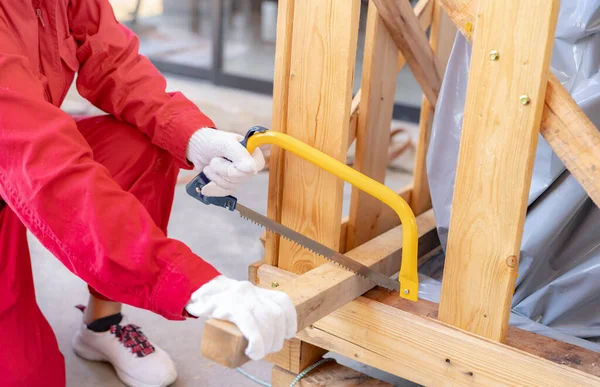 A worker is sawing wood to assemble a crate for moving an industrial machine. A woman in red mechanic coveralls hand holding a bow saw cutting the wooden plank of a crate. Safety in workplace concept.