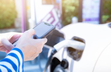 Woman using smartphone paying service in mobile app on blur EV car charging at electric vehicle charging station. EV car charging point. Commercial EV car charging station. Sustainability lifestyle.