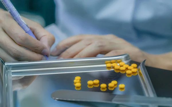 Stock image Yellow sugar coated tablet pill on stainless steel drug tray. Pharmacist hand holding pen and writing drug instructions on plastic pill pack. Prescription drug. World pharmacist day. Pharmacy concept.