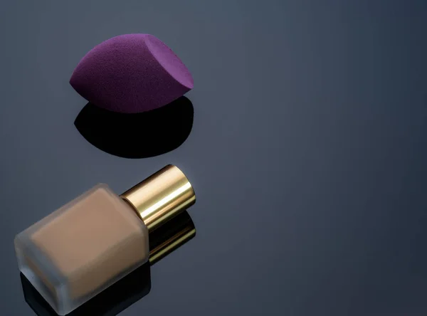 Purple makeup sponge on dark background. Soft makeup cosmetic sponge. Flat-ended makeup sponge with blur makeup foundation bottle. Liquid beige make-up foundation and beauty blender. Cosmetic product.