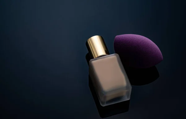 Purple makeup sponge on dark background. Soft makeup cosmetic sponge. Flat-ended makeup sponge with blur makeup foundation bottle. Liquid beige make-up foundation and beauty blender. Cosmetic product.