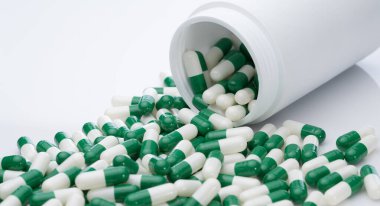 Beyaz plastik bir şişeden yeşil ve beyaz kapsül hapları döküldü. İlaç sanayisi. Reçeteli ilaç. Sağlık ve ilaç servisi. Eczane ürünü. Eczacılık. Kapsül hapı imalatı.