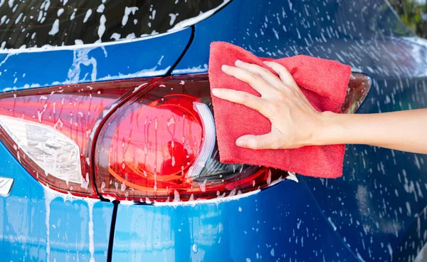 Car wash service. Car covered with white soap foam. Man hand holding red microfiber cloth and polish tail light of blue car. Auto care service business concept. Man cleaning and detailing luxury car.