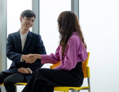 Bir adam mor gömlekli bir kadınla tokalaşıyor. Adam takım elbise ve kravat takıyor. Kadın sarı bir sandalyede oturuyor.