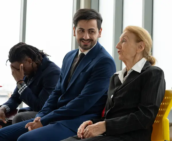 A man in a suit is sitting next to an older woman in a business suit. The man is smiling and the woman is looking at him