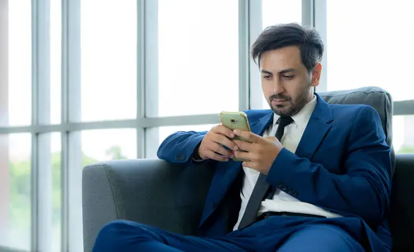 A man in a suit is sitting on a couch and looking at his cell phone. He is focused on the screen, possibly checking his messages or browsing the internet. Concept of modernity and technology