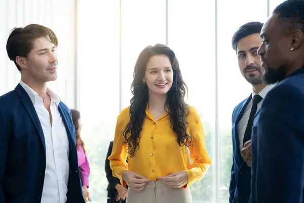 A group of people are standing in a room, one of them is wearing a yellow shirt. The woman in the yellow shirt is smiling and looking at the camera. The other people in the room are dressed in suits