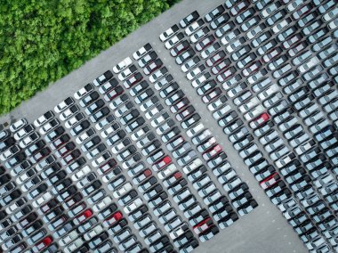 Yeşillik ormanın bitişiğindeki yeni arabalarla dolu bir fabrika otoparkının havadan görüntüsü. Otomotiv endüstrisi sürdürülebilirlik, lojistik ve sıfır emisyonlu elektrikli ve hibrit araçlara odaklanıyor.