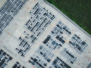 Yeşillik çim tarlasına bitişik yeni arabalarla dolu bir fabrika otoparkının havadan görüntüsü. Otomotiv endüstrisi sürdürülebilirlik, lojistik ve sıfır emisyonlu elektrikli ve hibrit araçlara odaklanıyor.