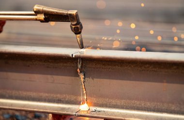 Kaynakçı metal kirişi kesmek için gaz lambası kullanıyor, kıvılcımlar uçuşuyor. Yetenekli işçilik ve endüstriyel metal üretiminde riskler. Hassasiyet ve işlem tehlikesi. Bir meşaleyle çelik kiriş kesiliyor
