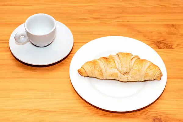 Baked croissant on ceramic plate with empty coffee cup on wooden table