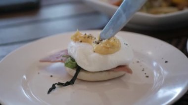 Beyaz tabakta yumurtalı Benedict yemek için bıçak ve çatal kullanarak yumurta sarısı kesiyor. Yumurta buğulama ve yumurta sarısı. Sağlıklı kahvaltı ya da öğle yemeği.