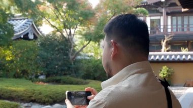 asian man enjoy relaxing breathe and take photo with smartphone in japanese zen style garden in autumn daytime, relax peaceful in japan garden style