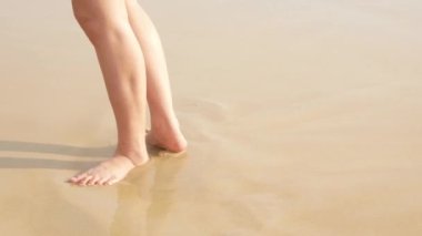 Düşük açı, çıplak ayakla sahile vuran deniz dalgalarıyla beyaz kumsalda hareket eden çıplak ayaklar, yaz tatili, doğal arka plan 4K. Kumsalda rahatça yürümek.