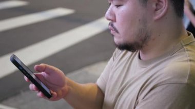 Asyalı bir adamın gece vakti alışveriş alanında Zebra geçişi geçmişiyle seyahat ederken akıllı cep telefonu mesajları kullandığı görüntüsü. Şehir caddesinde renkli bokeh ile mesajlaşma