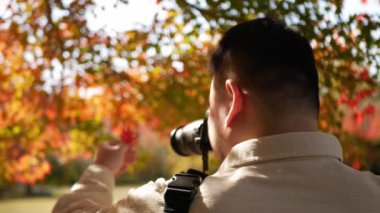 Asyalı adam akçaağaç yapraklarının arka planında renk değişikliği olan bir akçaağaç yaprağını tutarken fotoğraf çekiyor.