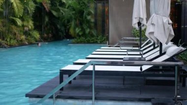 Güneş yatağı, Chaise Lounge ya da yüzme havuzu yanında temiz mavi suları ve modern tatil beldesini çevreleyen tropik ağaçları olan yüzme havuzu, yaz tatili arka planı.