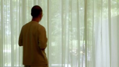 Elinde bir fincan kahve tutan Asyalı bir adam sabah beyaz şeffaf perdeden dışarı bakmak için yürüyor. Sağlıklı bir ferahlama için, yaz tatili sabahı