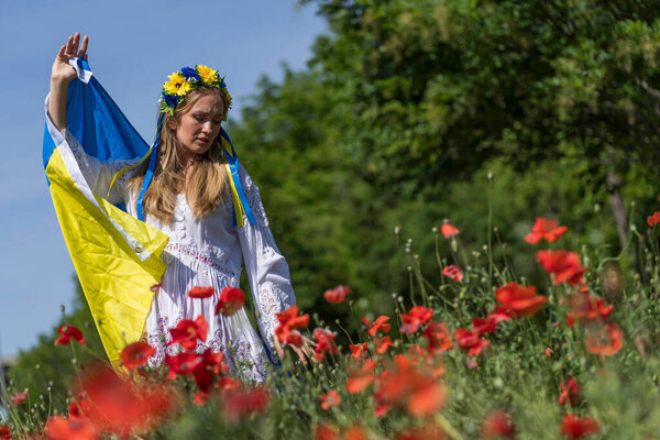 A young blonde Ukrainian woman stands in a field of Red Poppy flowers holding the flag of Ukraine showing her support for the war in her native country of Ukraine