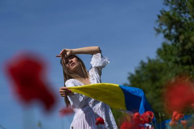 A young blonde Ukrainian woman stands in a field of Red Poppy flowers holding the flag of Ukraine showing her support for the war in her native country of Ukraine