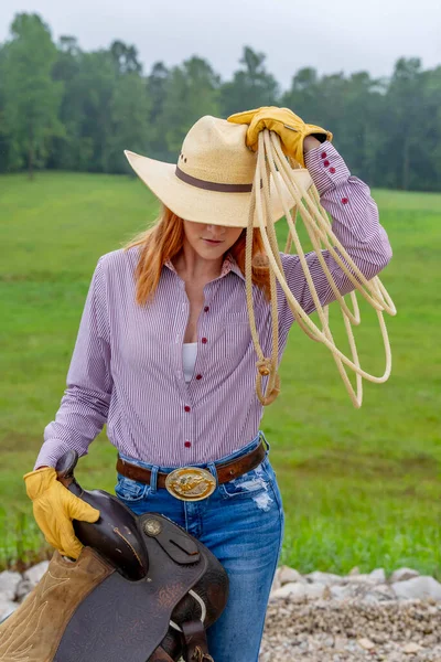 一个漂亮的红头发模特站在一个乡村 穿着牛仔女郎的服装 手握马鞍和绳索 — 图库照片