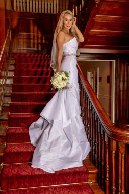 Gelinlik içindeki parlak sarışın gelin, düğün öncesi fotoğraflar için zarifçe poz veriyor, özel gününün heyecanını ve güzelliğini yakalıyor..