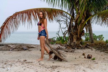 Bikinili, güneşli esmer, kumlu sahillerde Karayipler 'de eğleniyor, güzelliği macera ile birleştirerek hindistan cevizi avlıyor.