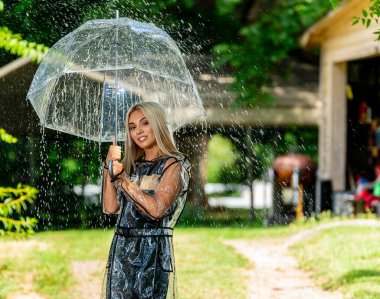 A radiant blonde model gracefully poses in the rain, donning a raincoat and holding an umbrella, her smile adding charm to the spring scene. clipart